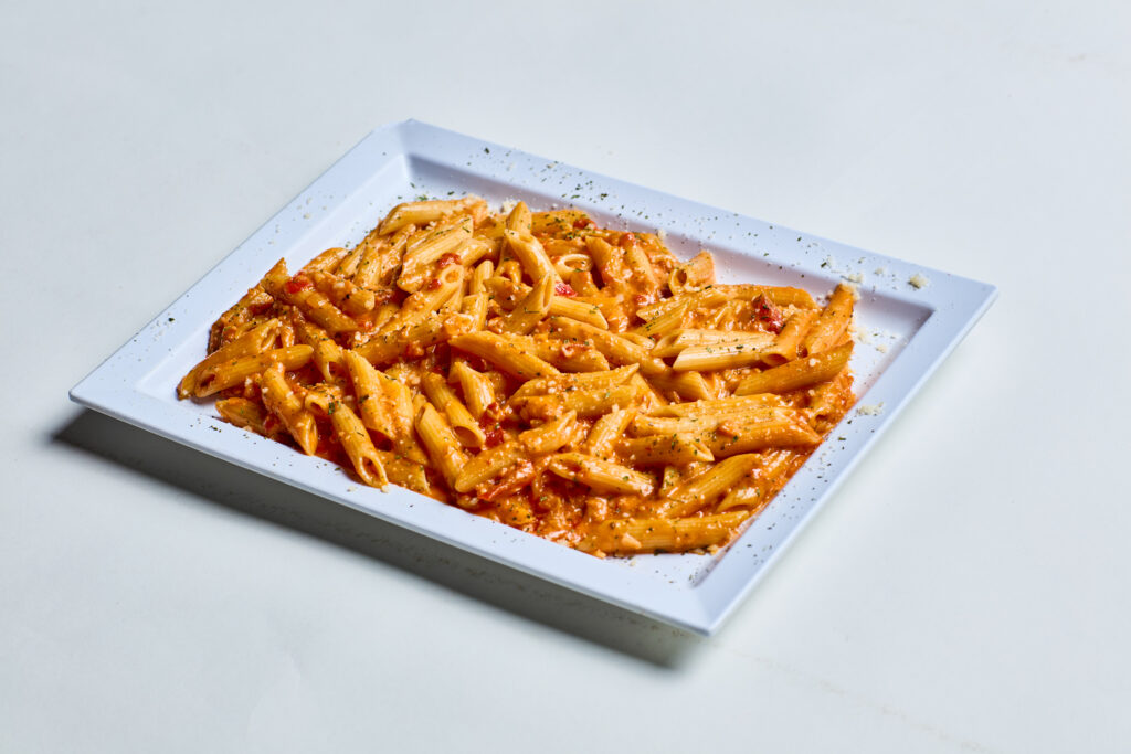 This image shows a plate of penne pasta coated in a creamy tomato sauce, garnished with grated cheese and herbs. The dish is served on a white rectangular plate against a light background.