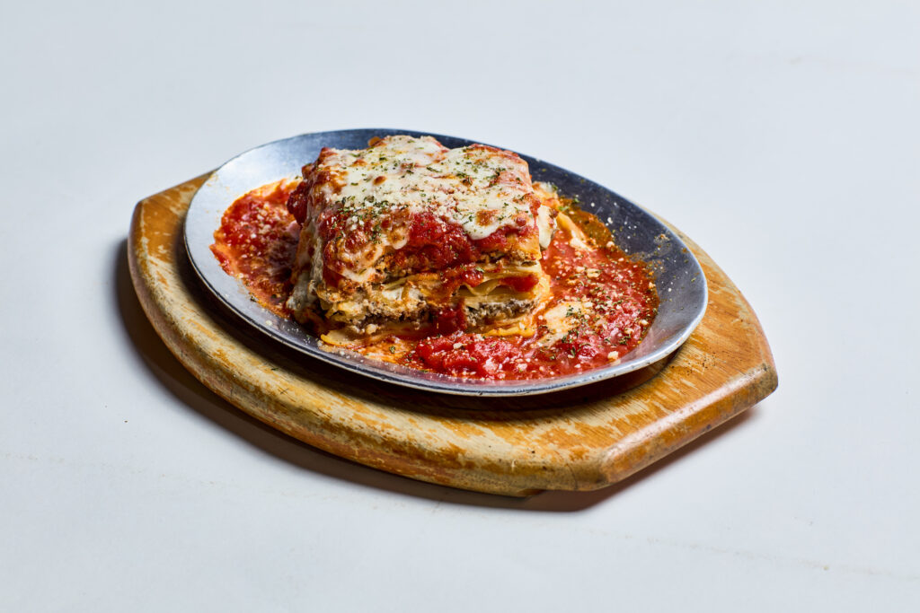 This image shows a serving of lasagna layered with pasta, meat sauce, ricotta, and melted cheese, topped with marinara sauce and sprinkled with herbs. It’s presented on a metal plate placed on a wooden serving board.
