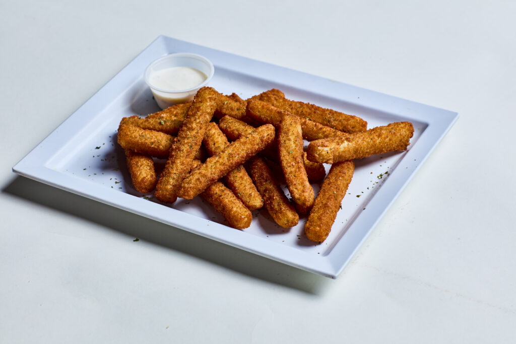 This image shows a plate of fried mozzarella sticks, golden brown and crispy, garnished lightly with herbs. A small cup of creamy dipping sauce, likely ranch or marinara, sits in the corner of the white rectangular plate.