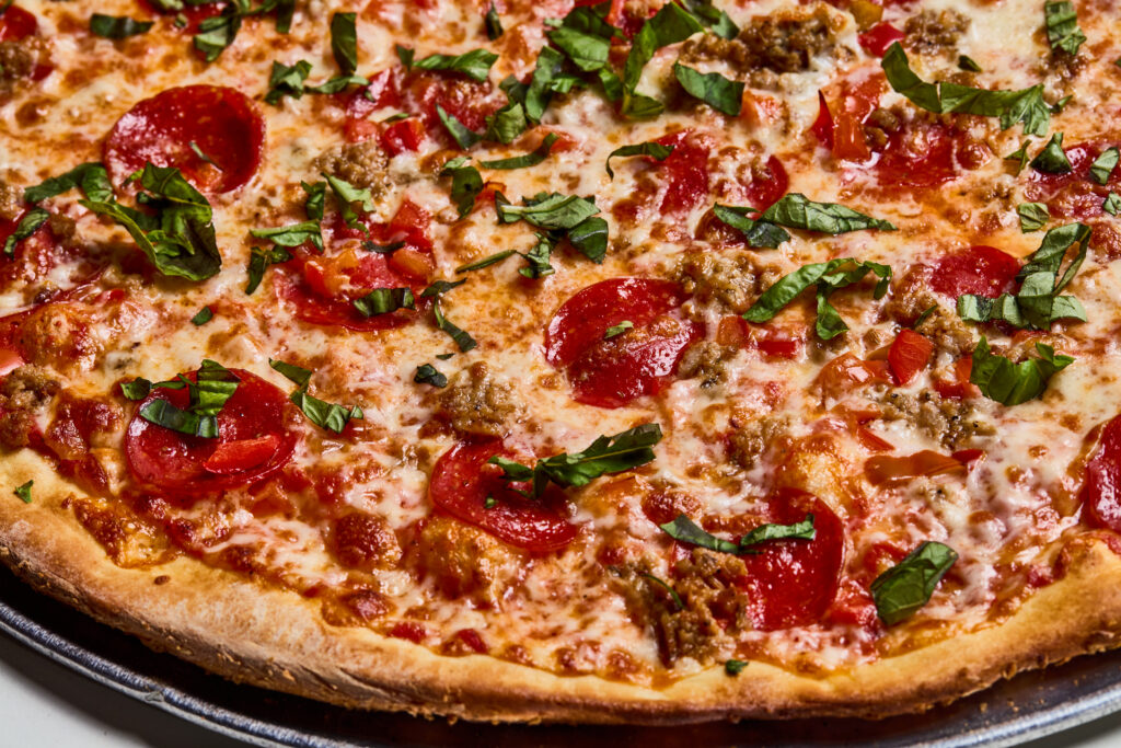 This image shows a freshly baked pizza topped with pepperoni, sausage, diced tomatoes, melted cheese, and chopped basil. The crust is golden brown and crisp, and the vibrant toppings stand out against the melted cheese base.