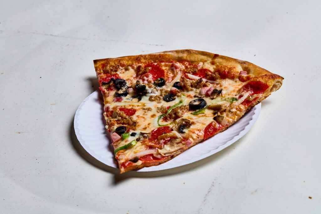 This image shows a slice of Supreme pizza on a white paper plate. The slice has toppings like pepperoni, sausage, ham, black olives, green peppers, onions, and melted cheese on a golden-brown crust, photographed on a light surface.