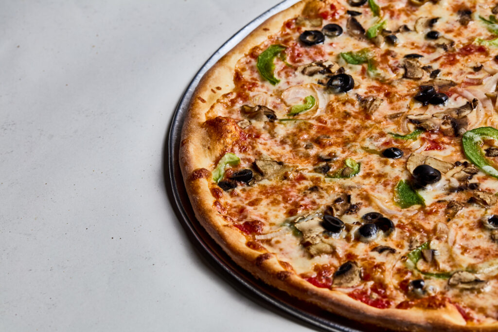 This image shows a vegetable pizza with a golden crust, topped with black olives, green bell peppers, onions, mushrooms, tomato sauce, and melted cheese. It sits on a metal pizza pan over a light gray surface, photographed from an angle that highlights its crisp edges and colorful toppings.