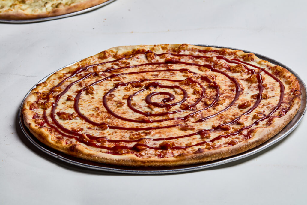 This image shows a barbecue chicken pizza with a spiral drizzle of barbecue sauce over a layer of melted cheese and chunks of chicken. The pizza has a golden-brown crust and is served on a metal pan.