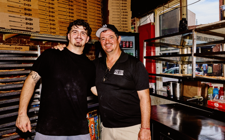 Two smiling pizza makers, one younger with a tattoo and the other older with a Yankees cap and a 'New York New York' shirt, stand together with arms around each other in a kitchen full of pizza boxes and metal racks.